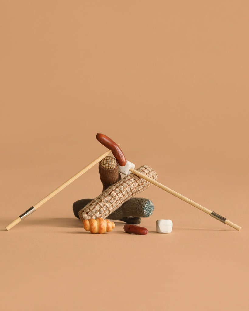 Two chopsticks form an upside-down V over log-shaped fabric, a plush croissant, and marshmallows—this whimsical Maileg Bonfire set inspires imaginative camping adventures, displayed on a plain beige background.
