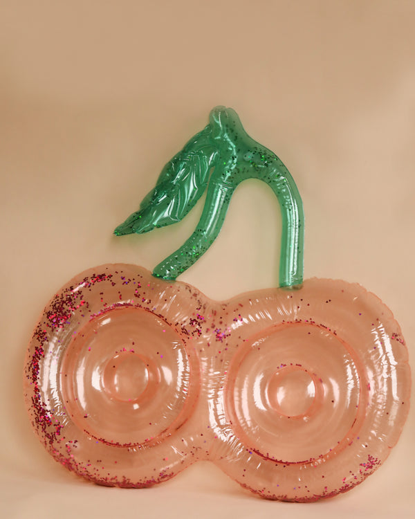 An Inflatable Cherry Float, resembling a playful toy, is set against a plain beige background. The cherry portion is transparent pink with embedded glitter, while the stem and leaf are transparent green. Ideal for shallow water, its two cherry circles are connected in the middle.