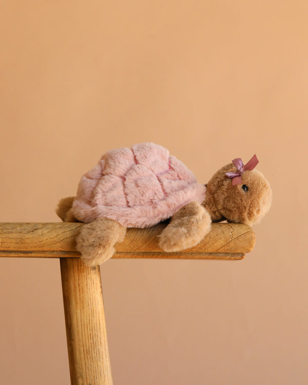 The Bukowski - Baby Sandy Sea Turtle - Pink plush, featuring a purple bow, sits on a wooden stool against a beige backdrop—an adorable pink stuffed animal perfect for any collection.