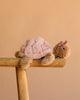 The Bukowski - Baby Sandy Sea Turtle - Pink plush, featuring a purple bow, sits on a wooden stool against a beige backdrop—an adorable pink stuffed animal perfect for any collection.
