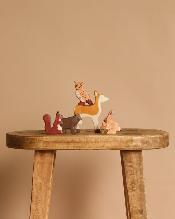 The Holztiger Handmade Wooden Woodland Animals Set, featuring a fox, bear, deer, owl, and squirrel, is displayed on a rustic stool against a plain beige background.