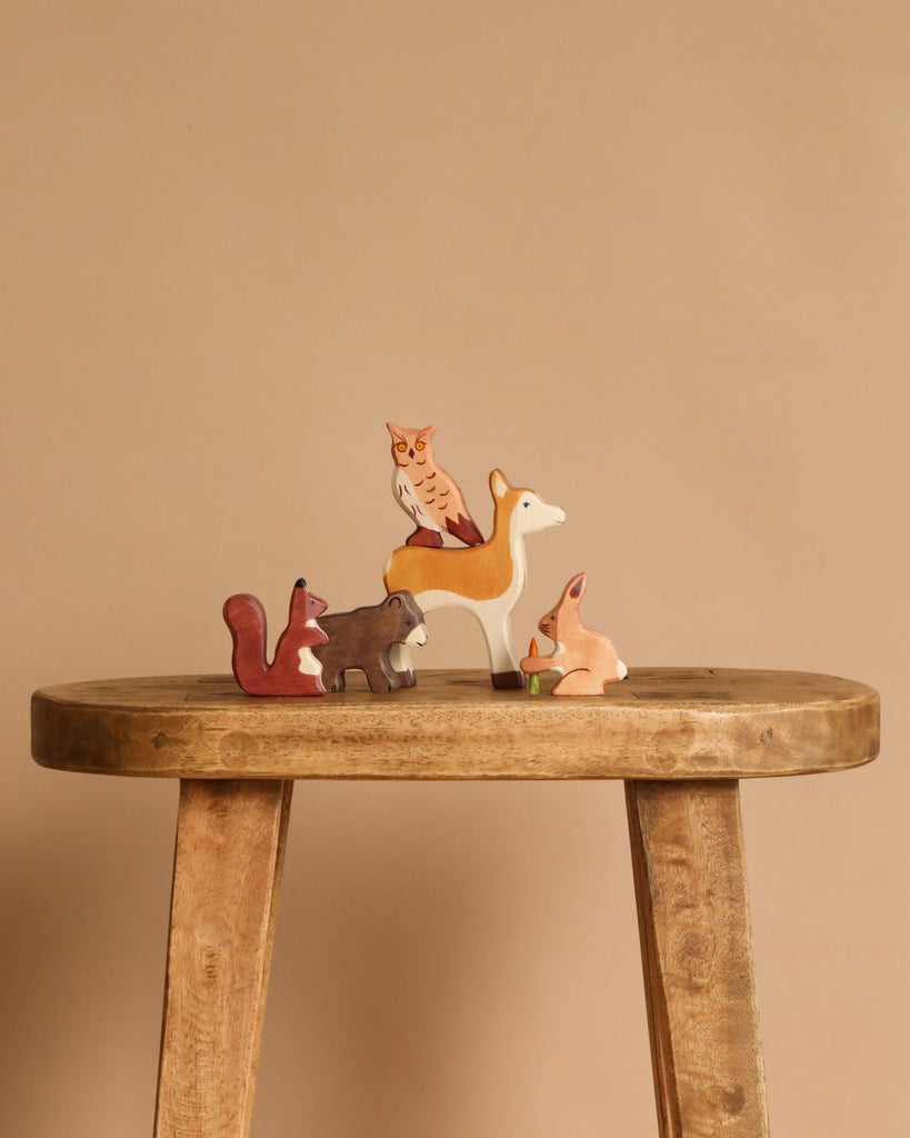 The Holztiger Handmade Wooden Woodland Animals Set, featuring a fox, bear, deer, owl, and squirrel, is displayed on a rustic stool against a plain beige background.