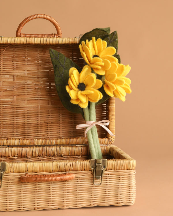 The Handcrafted Felt Flower Bouquet, featuring yellow flowers and green leaves tied with a white ribbon, is displayed in an open wicker basket against a beige background.