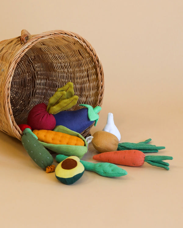 A basket tipped over, spilling the Handmade Soft Vegetable Set including carrots, a cucumber, corn, and an avocado, on a beige background.