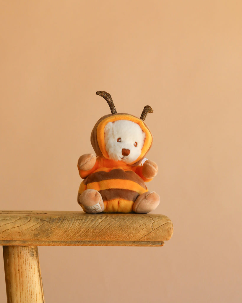 A Bukowski - Ziggy Bee plush toy, wearing an orange and brown bee costume with antennae, sits on the edge of a wooden stool against a plain beige background.