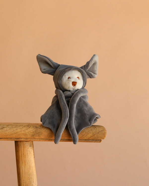 Bukowski - Ziggy Bat is a small white plush toy dressed in a gray hooded bat costume with large ears and soft velvet wings, shown sitting on a wooden stool against a beige background. Ideal as a charming Halloween plush.