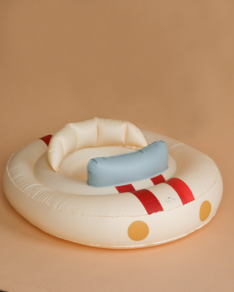 A round, cream-colored inflatable swim ring designed like a boat with red stripes and two yellow circles on the front. It features a backrest with a blue and white cushion attached, sitting on a light brown surface with a beige background. Ideal for children aged 1-2 years, made from durable PVC. Introducing the Inflatable Baby Swim Ring - Car!