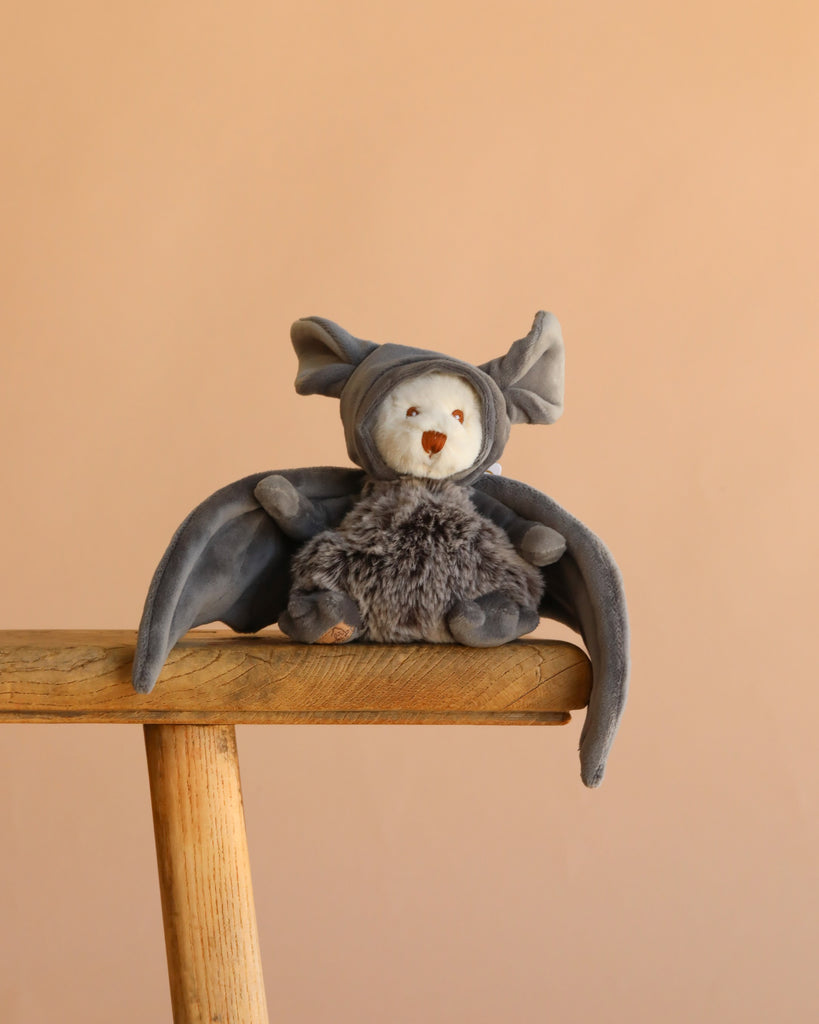 Bukowski - Ziggy Bat, a gray Halloween plush with soft velvet wings and pointed ears, sits on a wooden stool against a plain beige background.