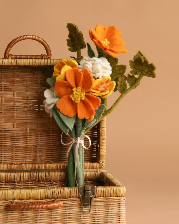 The Handcrafted Felt Flower Bouquet features orange, yellow, and white felt flowers with green leaves, tied with a pale ribbon and displayed in an open woven wicker suitcase against a beige background.