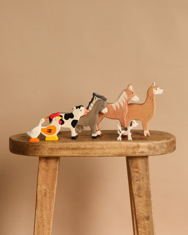 Six charming figures from the Holztiger Handmade Wooden Farm Animals Set—a duck, chicken, cow, donkey, horse, and dog—are arranged in a row on a wooden stool against a plain beige background.