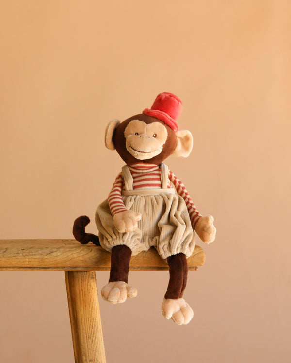 Bukowski - Winston Monkey - Tan, a plush toy in a red hat, striped shirt, and beige overalls, sits atop a wooden stool against a simple beige background.