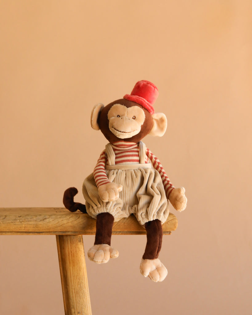 Bukowski - Winston Monkey - Tan, a plush toy in a red hat, striped shirt, and beige overalls, sits atop a wooden stool against a simple beige background.