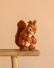 The Steiff Niki Squirrel, featuring brown fur, a fluffy tail, and the classic Button in Ear, perches on a wooden bench against a plain beige background.