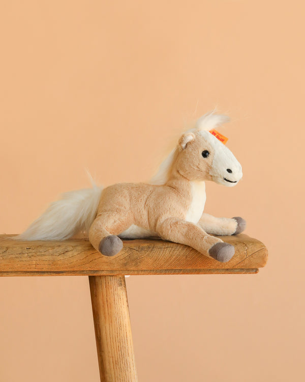 The Steiff Gola Dangling Horse, a light brown stuffed animal with a white mane and tail, sits on a rustic wooden bench against a plain peach background.