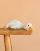 The Steiff Gilda Goose Rattle, a white plush toy with a yellow beak and feet, lies belly-down on a rustic wooden stool against a beige background—an adorable baby gift idea.