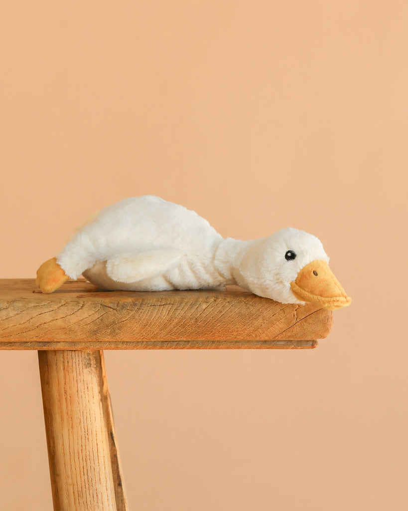 The Steiff Gilda Goose Rattle, a white plush toy with a yellow beak and feet, lies belly-down on a rustic wooden stool against a beige background—an adorable baby gift idea.