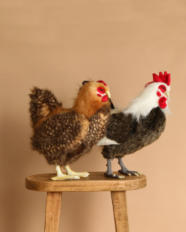 The Barnyard Duo Rooster and Hen Stuffed Animal Set features two hand-sewn plush chickens—one brown and speckled, the other dark brown with white neck feathers—displayed side by side on a wooden stool against a beige background.