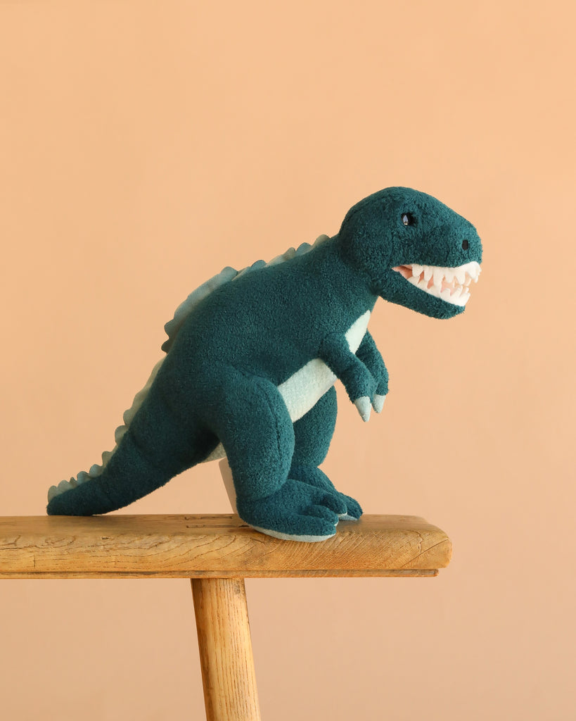 The Steiff Thaisen T-Rex Dinosaur, 14", a soft teal-blue plush with white teeth and light blue accents, sits on a wooden bench against a peach-colored background.