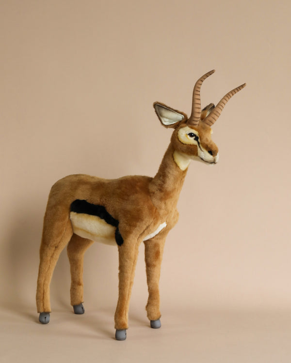 A realistic plush model of a Gazelle Stuffed Animal - 28" with prominent curved horns and distinct black markings on its sides, standing against a light beige background. This hand-sewn plush animal showcases artisan-crafted toys with realistic characteristics.