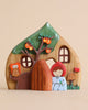 The Handmade Wooden Fairy Cottage is a delightful set that includes a charming small house with windows, a tree adorned with orange fruit, and a character dressed in red and wearing a white apron. The house's roof is green, mimicking foliage, while additional tiny details like a log and a small wooden door complete the enchanting ensemble.