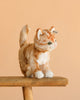 The Steiff Mizzy Cat, a realistic orange and white plush toy with green eyes, stands on a wooden bench against a plain peach background.