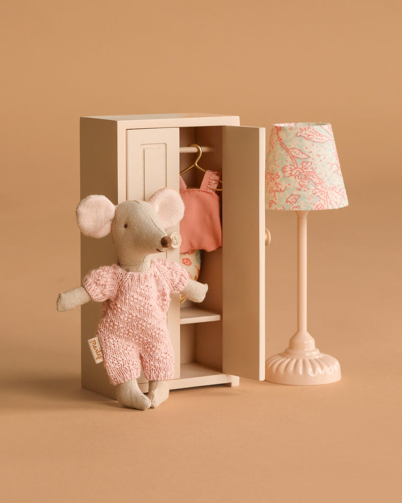 A Maileg Springtime Wardrobe Set features a small beach mouse in pink overalls beside a mouse closet filled with pink clothes and a cream vintage floor lamp with a floral shade, all set against a beige background.