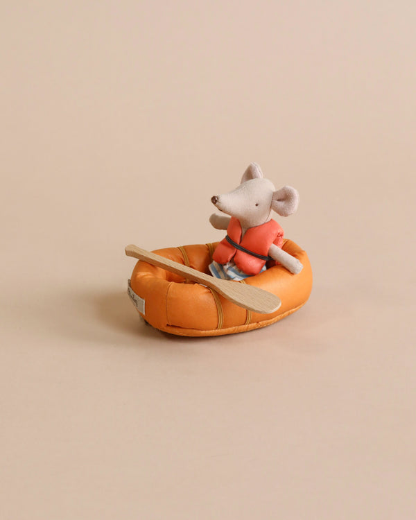 A small stuffed mouse wearing an orange life vest sits in a Maileg Rubber Boat, holding a wooden paddle, ready for a pretend water adventure. The boat is placed on a plain, light-colored background.