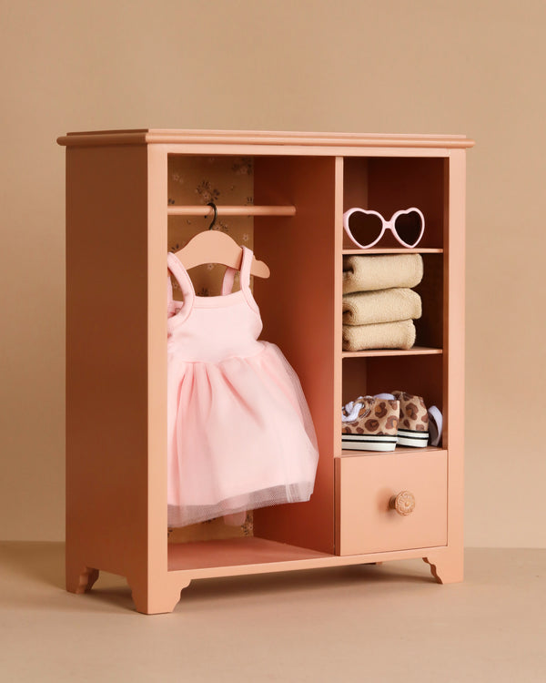 The Maileg Wardrobe, Large - Dark Powder (compatible with Minikane Dolls), holds a pink tutu dress, bunny and teddy outfits, folded towels, leopard-print clothes, heart-shaped sunglasses, and a small drawer—set against a beige background.
