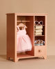 The Maileg Wardrobe, Large - Dark Powder (compatible with Minikane Dolls), holds a pink tutu dress, bunny and teddy outfits, folded towels, leopard-print clothes, heart-shaped sunglasses, and a small drawer—set against a beige background.