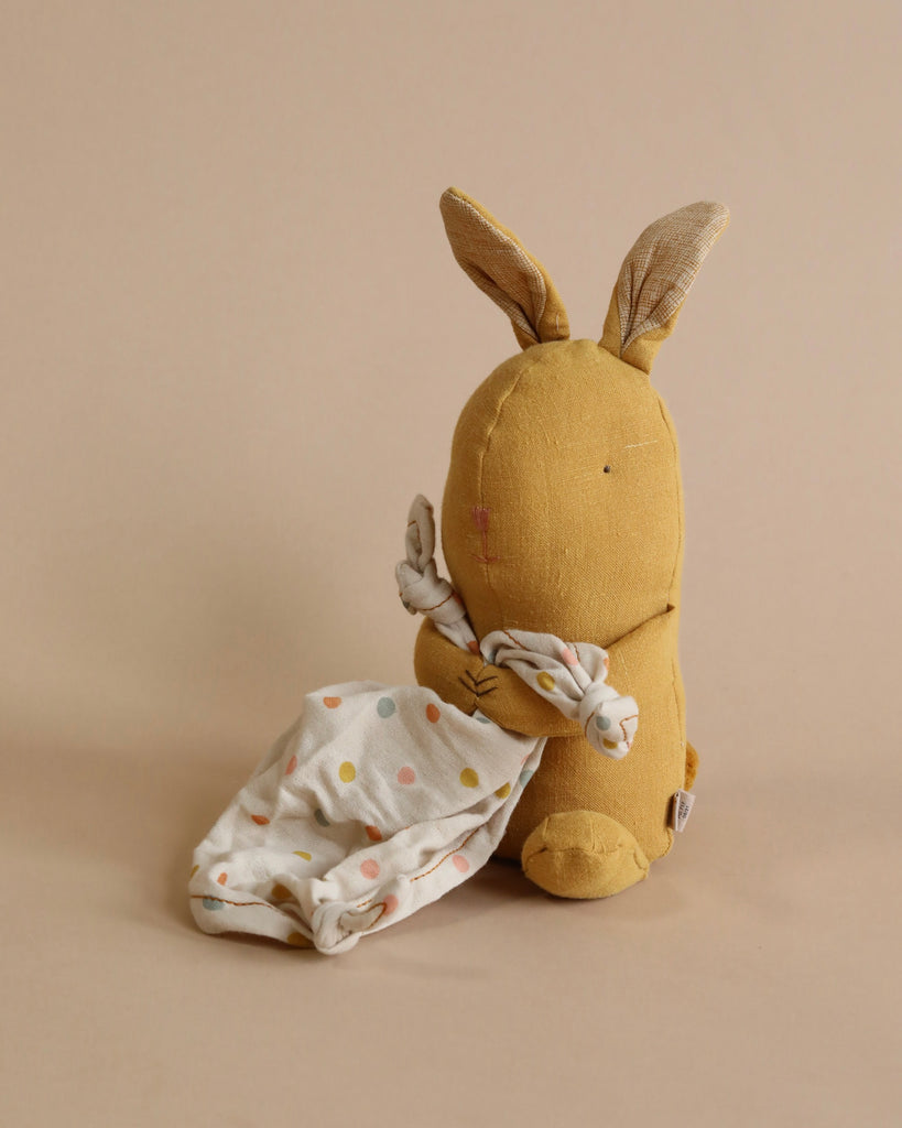 A Maileg Lullaby Friends - Bunny (Plays Music) with long ears, light brown color, and facing away from the camera is holding a white cloth with colorful polka dots. The background is a muted beige. This charming gift for babies brings a touch of whimsy to any nursery.