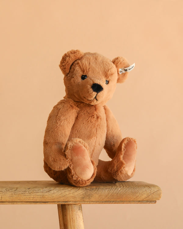 The Steiff Lio Teddy Bear, complete with an ear tag, sits upright on a wooden bench against a plain beige background.