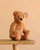 The Steiff Lio Teddy Bear, complete with an ear tag, sits upright on a wooden bench against a plain beige background.