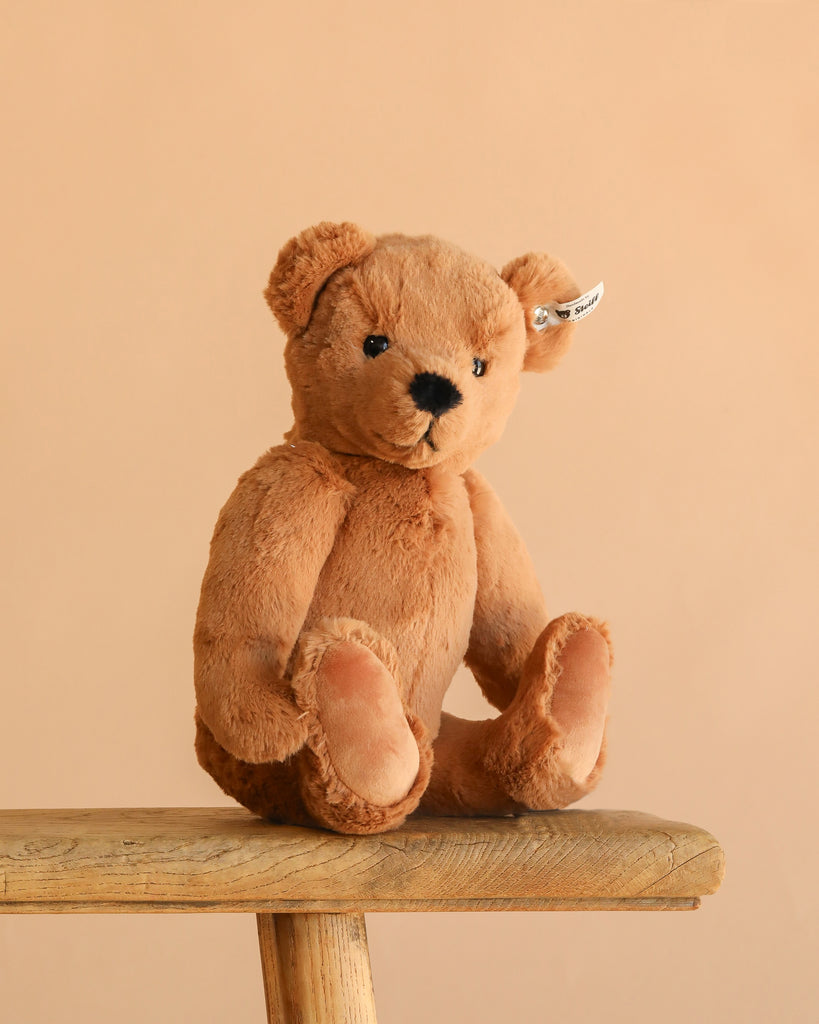 The Steiff Lio Teddy Bear, complete with an ear tag, sits upright on a wooden bench against a plain beige background.