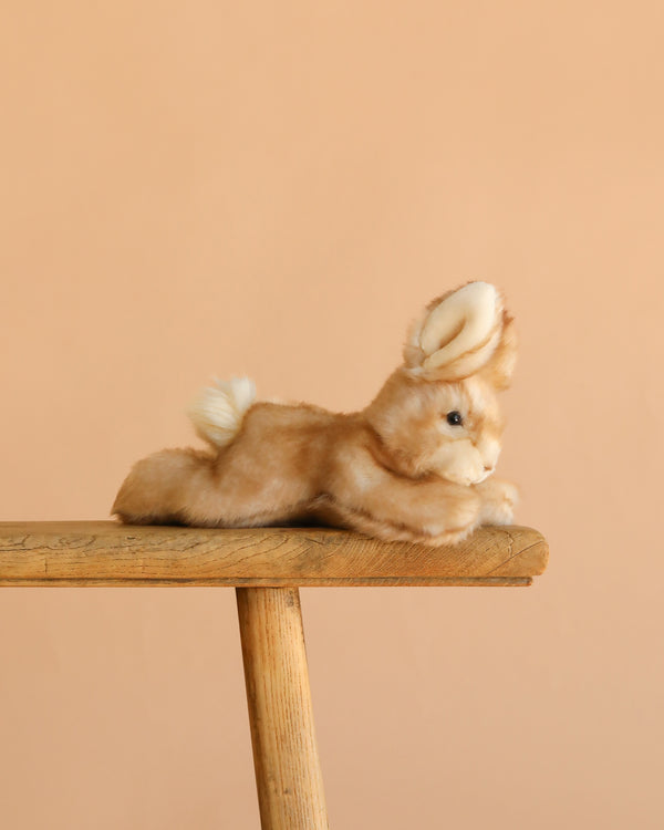 The Steiff Schnucki Rabbit, a hand-crafted plush in light brown with white accents, is shown stretched out on a wooden stool against a plain peach background.