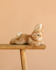 The Steiff Schnucki Rabbit, a hand-crafted plush in light brown with white accents, is shown stretched out on a wooden stool against a plain peach background.