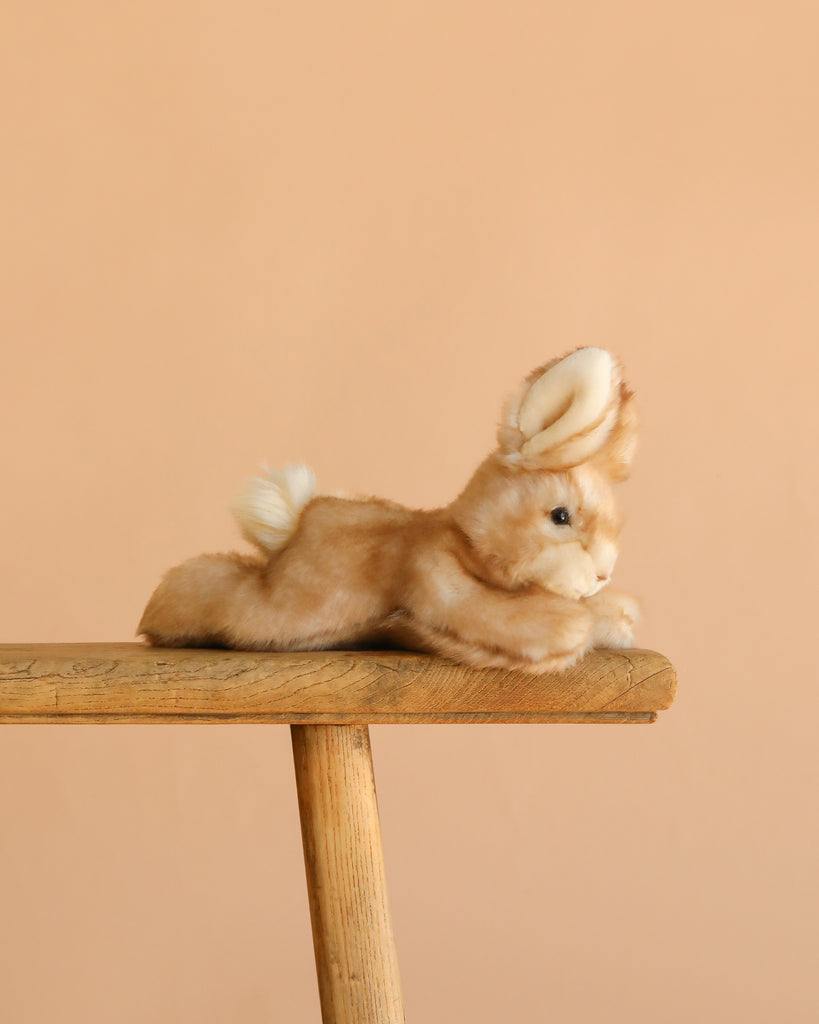 The Steiff Schnucki Rabbit, a hand-crafted plush in light brown with white accents, is shown stretched out on a wooden stool against a plain peach background.
