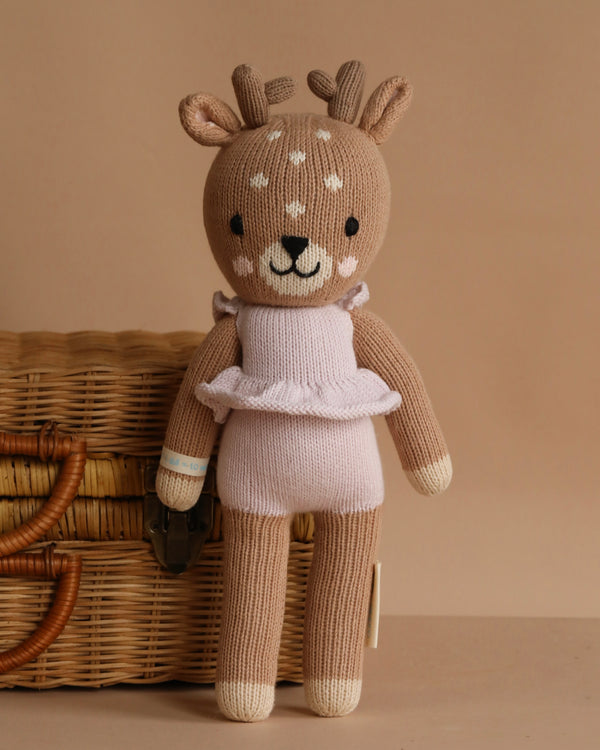 Cuddle + Kind Violet the Fawn is a hand-knit deer plush with antlers and white spots, dressed in a light pink ruffled outfit, made from premium cotton yarn, shown standing before a wicker basket on a beige background.