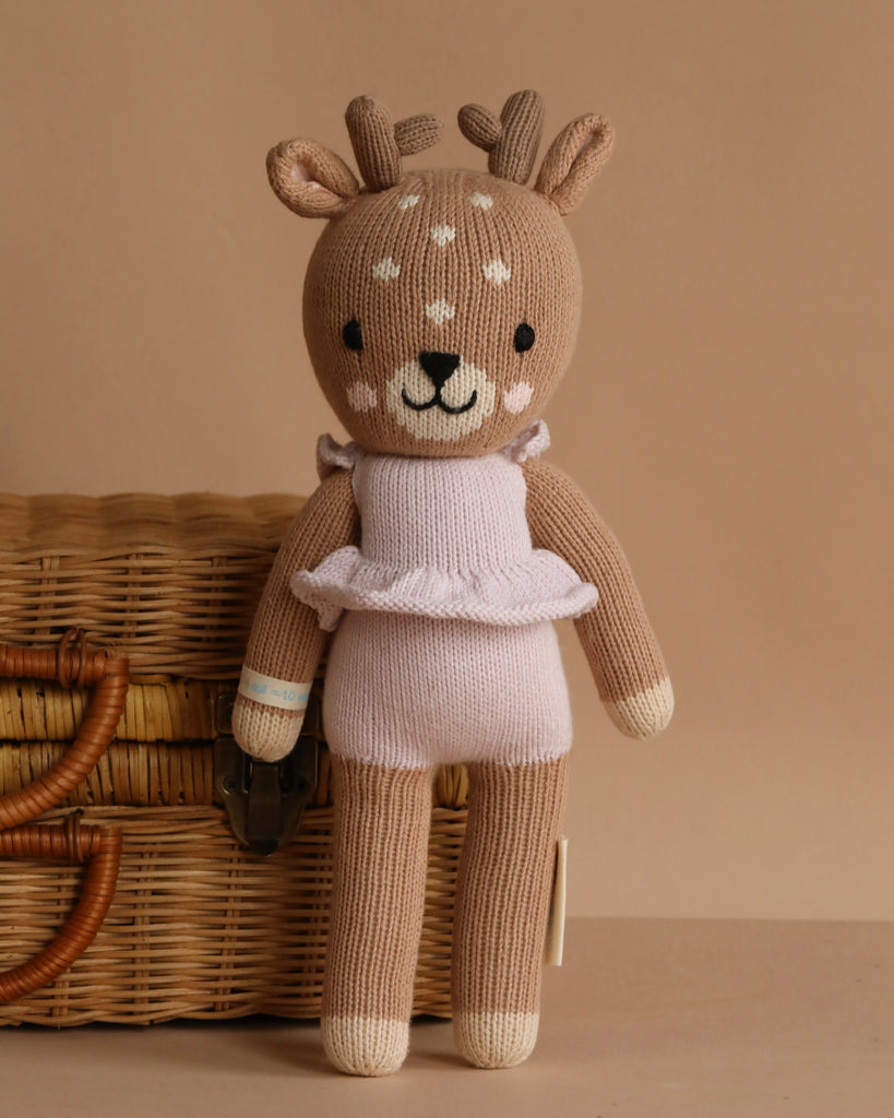 Cuddle + Kind Violet the Fawn is a hand-knit deer plush with antlers and white spots, dressed in a light pink ruffled outfit, made from premium cotton yarn, shown standing before a wicker basket on a beige background.