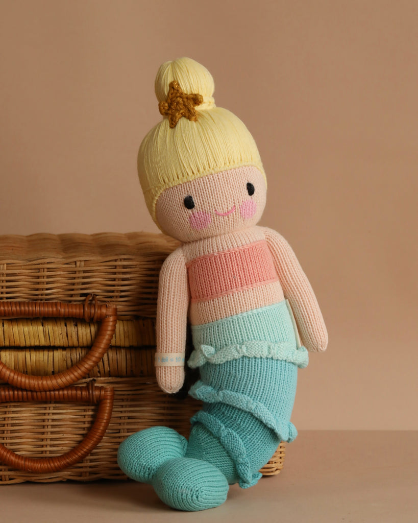 The fair trade handmade Cuddle + Kind Skye the Mermaid doll, with blonde hair in a bun, a starfish accessory, and a pastel tail, sits against a woven picnic basket on a warm beige background.