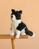 The Steiff Jaycee Border Collie Dog, a black and white stuffed toy, sits upright on a wooden stool against a plain beige background.