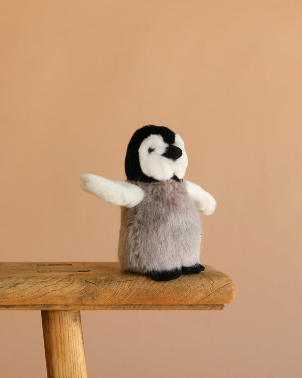 The Steiff Flaps Penguin, a small plush toy with outstretched wings, stands on a wooden stool against a plain beige background—an adorable heirloom piece for any collection.
