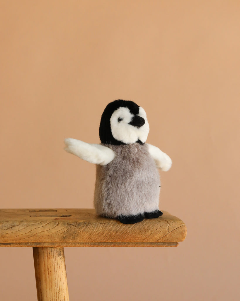 The Steiff Flaps Penguin, a small plush toy with outstretched wings, stands on a wooden stool against a plain beige background—an adorable heirloom piece for any collection.