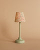 A small, pale green floor lamp from the Maileg Bedroom Set features a patterned red and cream lampshade and is displayed against a plain beige background.