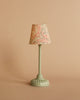 A small, pale green table lamp with a patterned fabric lampshade stands on a plain beige surface against a beige background, perfectly complementing the charm of the Maileg Cozy Corner Set and adding a touch of whimsy to your miniature scene.