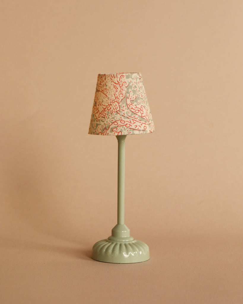 A small, pale green table lamp with a patterned fabric lampshade stands on a plain beige surface against a beige background, perfectly complementing the charm of the Maileg Cozy Corner Set and adding a touch of whimsy to your miniature scene.
