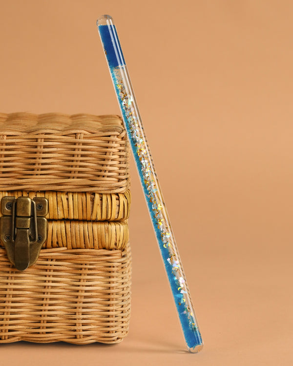 The Konges Sloejd Magical Glitter Wand - Rocket, a clear pen with blue ink and gold glitter, leans against a closed woven wicker box with a metal latch, set against a plain beige background.