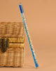 The Konges Sloejd Magical Glitter Wand - Rocket, a clear pen with blue ink and gold glitter, leans against a closed woven wicker box with a metal latch, set against a plain beige background.