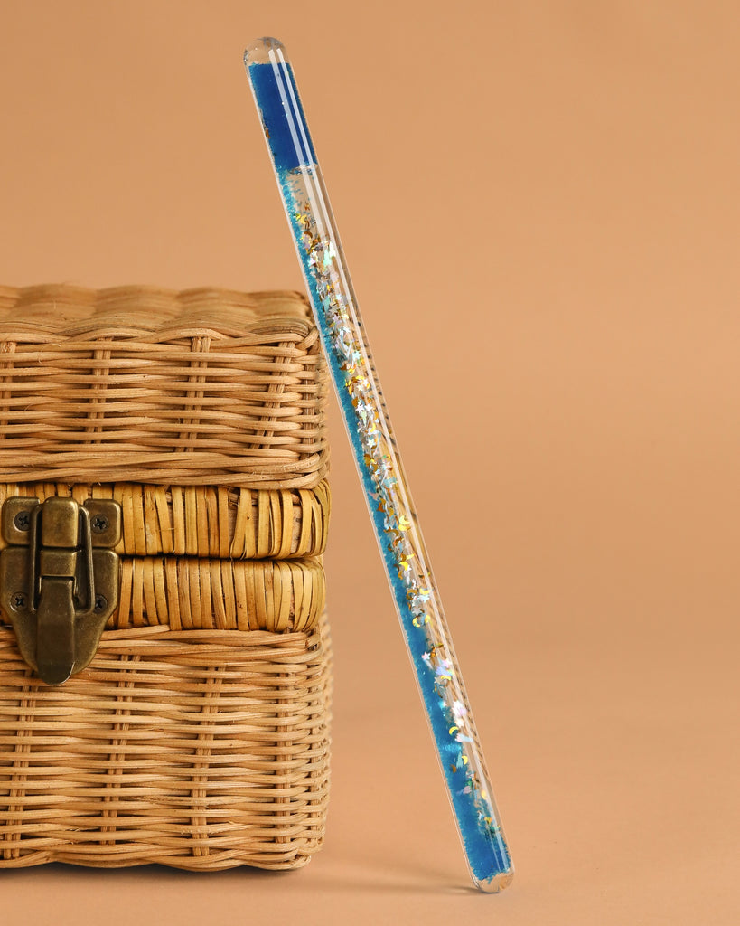 The Konges Sloejd Magical Glitter Wand - Rocket, a clear pen with blue ink and gold glitter, leans against a closed woven wicker box with a metal latch, set against a plain beige background.