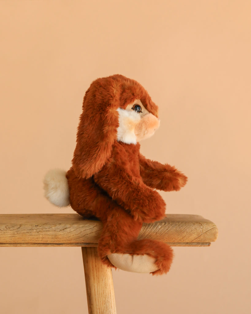 The Steiff Bommel Dangling Rabbit, a brown and white bunny plush with floppy ears, sits on a wooden stool against a beige background—ready to be your perfect snuggle buddy.