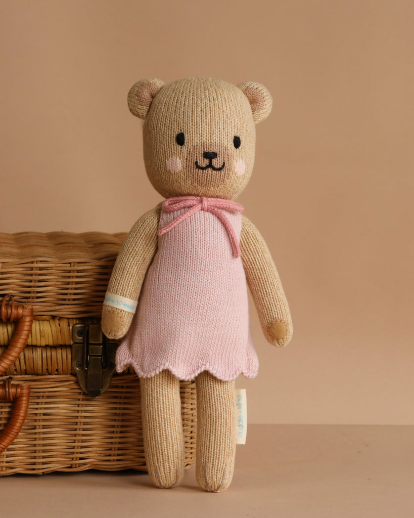 Cuddle + Kind Bella the Honey Bear, a hand-knit teddy bear in a pink dress, stands before a woven basket with a leather latch against a beige background.